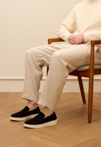 Black suede slip-on shoes with a white rubber sole, worn with light beige linen pants, seated on a wooden chair over a herringbone floor.