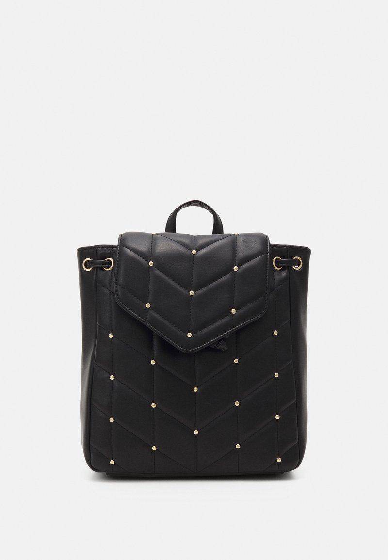 Sac à dos matelassé noir avec des clous dorés, fermeture à rabat, côtés à cordon de serrage et poignée supérieure sur fond gris clair uni.