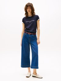 T-shirt bleu marine avec bordures blanches et texte "TOMMY HILFIGER", associé à un jean large bleu et des chaussures beiges à enfiler avec un accent métallique.