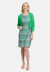 Grüner, kurz geschnittener Cardigan über einem multicolor gestreiften Kleid in Blau- und Grüntönen, mit knielangem Saum und weißen Sandalen.