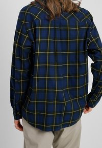 Camicia da uomo in flanella, blu scuro con motivo a quadri gialli e verdi, maniche lunghe, chiusura con bottoni, tessuto morbido e dettaglio sulla spalla posteriore.