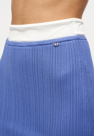 Vue sur le buste d'une personne portant un sous-vêtement blanc et une jupe côtelée bleue taille haute en maille, ornée d'une petite broche ovale argentée.