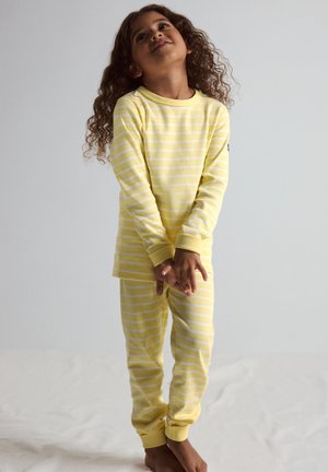 Jong meisje met krullend haar staat op blote voeten, draagt een geel-wit gestreept shirt met lange mouwen en een broek, kijkt omhoog met gevouwen handen.