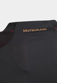 Mustaksi rakenteeltaan kankaalla on "DEUTSCHLAND" tulostettuna kullalla kauluksen lähellä, ja sivuilla on hienovaraisia punaisia ompeleita.