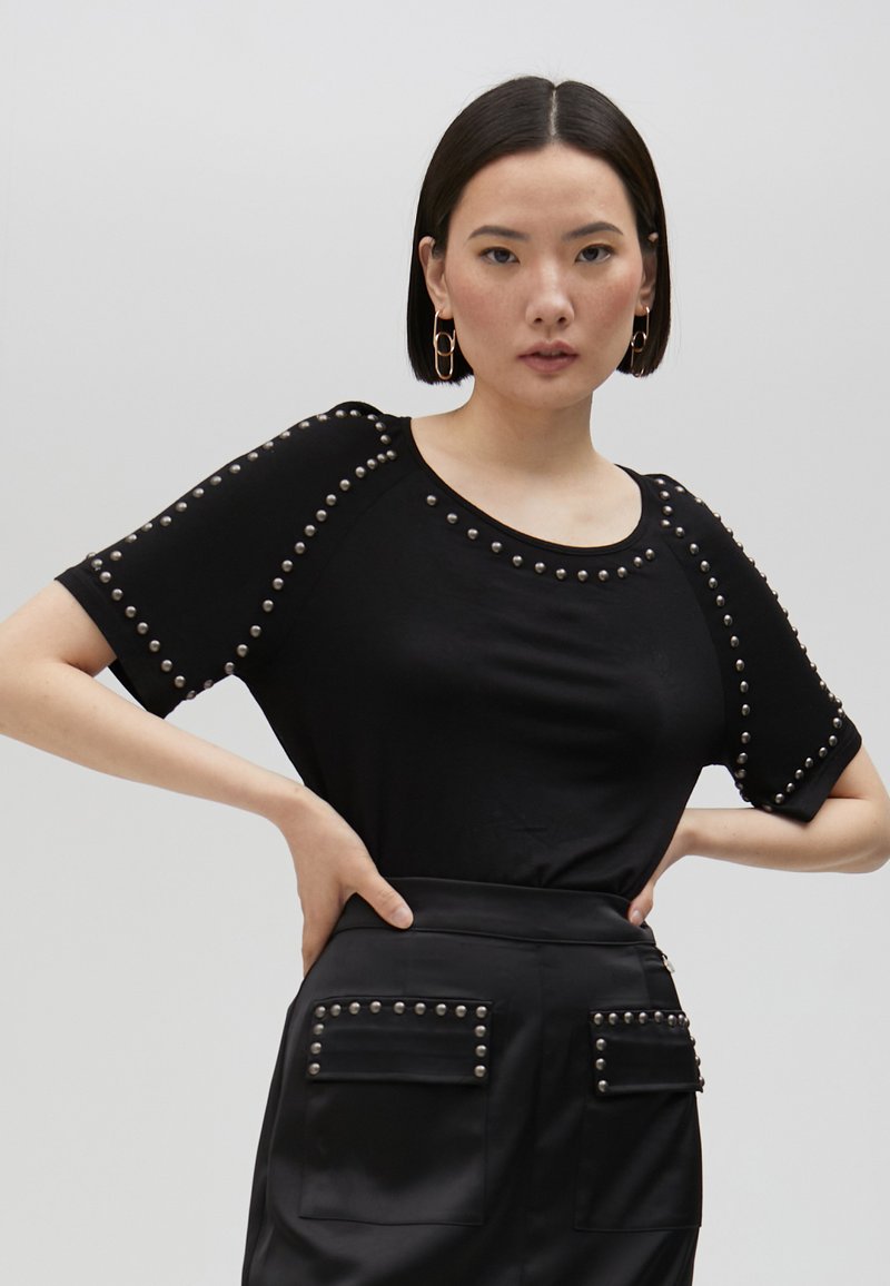 Černý top s krátkým rukávem a hroty podél rukávů a výstřihu, s volným střihem. V kombinaci s černými kalhotami s vysokým pasem a detaily s hroty.