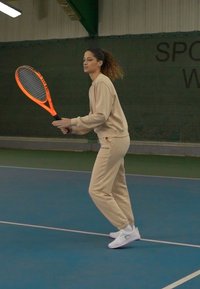 Beige sportoutfit met lange mouwen en broek, witte sportschoenen, met een oranje tennisracket in de hand op een binnenbaan met groene netten.