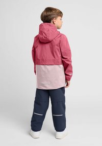Junge in einer zweifarbigen, rosa Kapuzenjacke und navyblauen Hosen mit reflektierenden Streifen, steht seitlich vor einem einfarbigen Hintergrund.