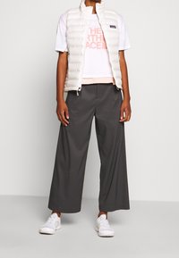 Osoba v bílé prošívané vestě přes světle růžové tričko The North Face, v širokých šedých kalhotách a bílých teniskách, stojící na šedém pozadí.