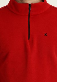 Pullover in pile rosso con colletto alto, zip frontale e logo nero accentato sul petto. Texture morbida, design minimalista.
