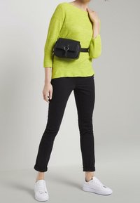 Haut en maille vert citron, jean skinny noir et baskets blanches. Un sac-ceinture noir avec un accent en métal rond porté à la taille.