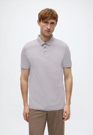 Ανοιχτό γκρι πόλο μπλούζα με γιακά, τέσσερα κουμπιά και κοντά μανίκια. Το ύφασμα φαίνεται λείο με ελαφριά γυαλάδα, σε συνδυασμό με μπεζ παντελόνι.