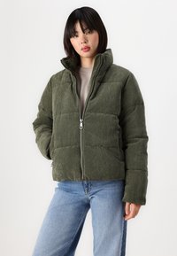 Olivengroene corduroy pufferjack met een hoge kraag, ritssluiting en een gewatteerd patroon, gedragen met lichtblauwe jeans.