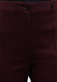 Gros plan sur un pantalon bordeaux avec un bouton et une ceinture visibles, présentant des coutures minimales et une texture de tissu lisse.