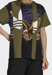 Olivgrön och svart kortärmad t-shirt med en stor vit logotyp och en örn grafik mot en mörk, molnig bakgrund.