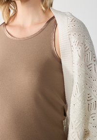 Tělová žebrovaná tílko v kombinaci s bílým otevřeným pleteným kardiganem s ozdobnými vzory podél okrajů.