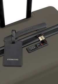 Olijfgroene harde koffer met een zwarte uitschuifbare handgreep, dubbele ritsen, een zwart bagagelabel en een paneel voor een cijferslot.