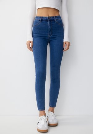 Femme portant un jean skinny bleu taille haute, un crop top blanc et des baskets blanches, debout devant un fond clair uni.