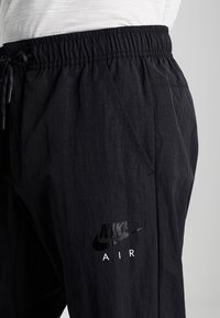Černé kalhoty Nike Air na stažení, nošené s bílým tričkem, ukazující elastický pas a detaily bočních kapes.