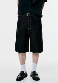 Férfi fekete denim térdhoszú sortot, fekete övet, fehér sportzoknit és fekete bőr loafert visel, fehér háttér előtt áll.