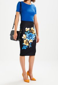 Top blu a maniche corte, gonna a matita con motivo floreale nera con fiori gialli e blu, tacchi gialli e una borsa nera.