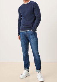 Homme portant un pull bleu marine, un jean bleu retroussé, des baskets blanches et un sous-pull blanc, debout avec une main dans la poche.