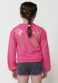 Sweatshirt rose avec des motifs d'ailes dorées dans le dos, associé à un short foncé. Le sweatshirt a une coupe décontractée et des manches longues.
