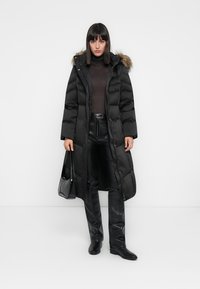 Svart lang puffjakke med hette fra pels, med kviltet design, frontknapplukking, kombinert med svarte skinnbukser og hælboots.