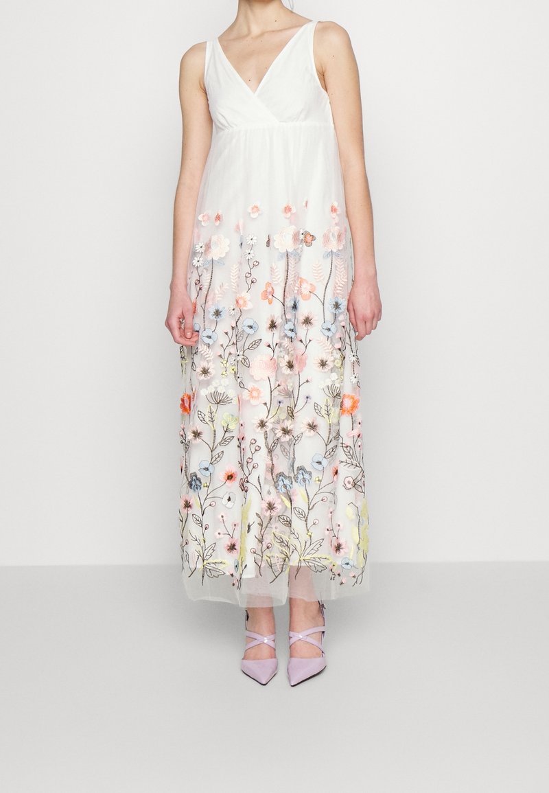 Femme portant une robe maxi blanche sans manches avec une broderie florale colorée et des talons pointus lavande sur fond uni.