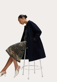 Cappotto blu navy con bottoni dorati, indossato sopra un vestito a stampa leopardata. Modella seduta su uno sgabello bianco, che espone tacchi a punta bianchi.