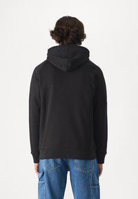 Felpa nera con cappuccio dal tocco morbido, con polsini e orlo a coste, caratterizzata da maniche a raglan per un fit rilassato. Indossata con pantaloni di denim blu chiaro.