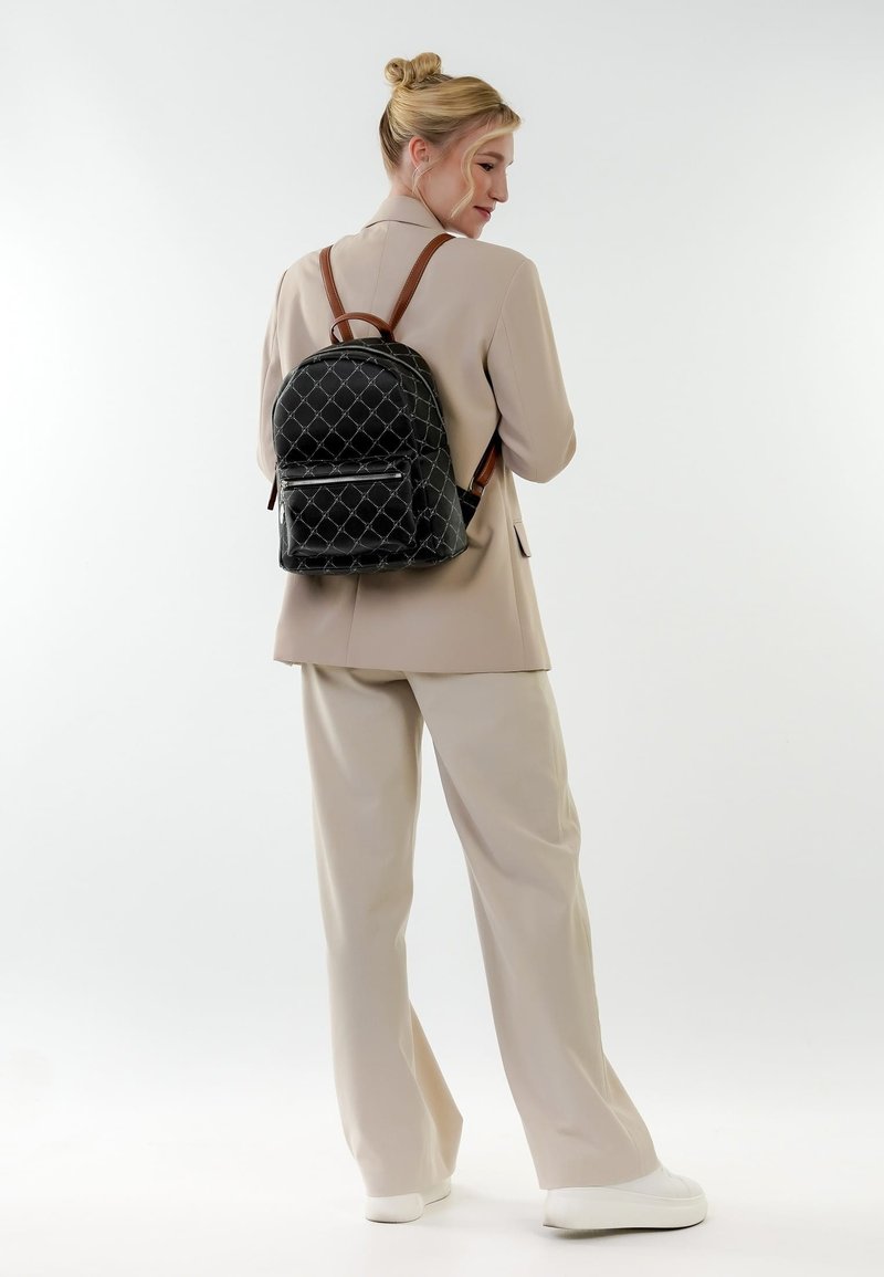 Vaaleatukkainen nainen nutturalla, pukeutunut beigeen pukuun ja valkoisiin tennareihin, kantaa selässään mustaa kuviollista reppua ja katsoo olkansa yli.