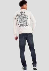 Sudadera color crema con texto en la espalda: "Preferiría estar cansado que en quiebra." Combinada con jeans de mezclilla oscura y zapatillas negras.