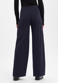 Vide bukser i mørk marineblå, med et glatt stoff, høy midje og subtile sømdetaljer, kombinert med svarte platåsko.