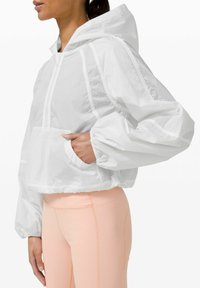 Kratka bela vetrovka s kapuco, elastičnimi manšetami in žepom; lahka, teksturirana tkanina je vidna, v kombinaciji s svetlo breskevnimi leggins.