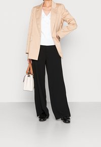 Blazer beige sopra una blusa bianca con volant e pantaloni neri a gamba larga. Le cinghie di una piccola borsa bianca abbinano le accentuazioni marroni; le scarpe nere completano il look.