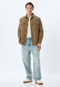 Chaqueta de camisa de pana marrón con bolsillos en el pecho abotonados, llevada sobre una camiseta clara, combinada con unos jeans de corte holgado en azul claro y zapatillas blancas.
