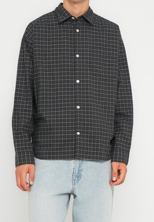 Mand iført sort ternet skjorte med lange ærmer med hvide linjer og lyseblå jeans mod hvid baggrund.