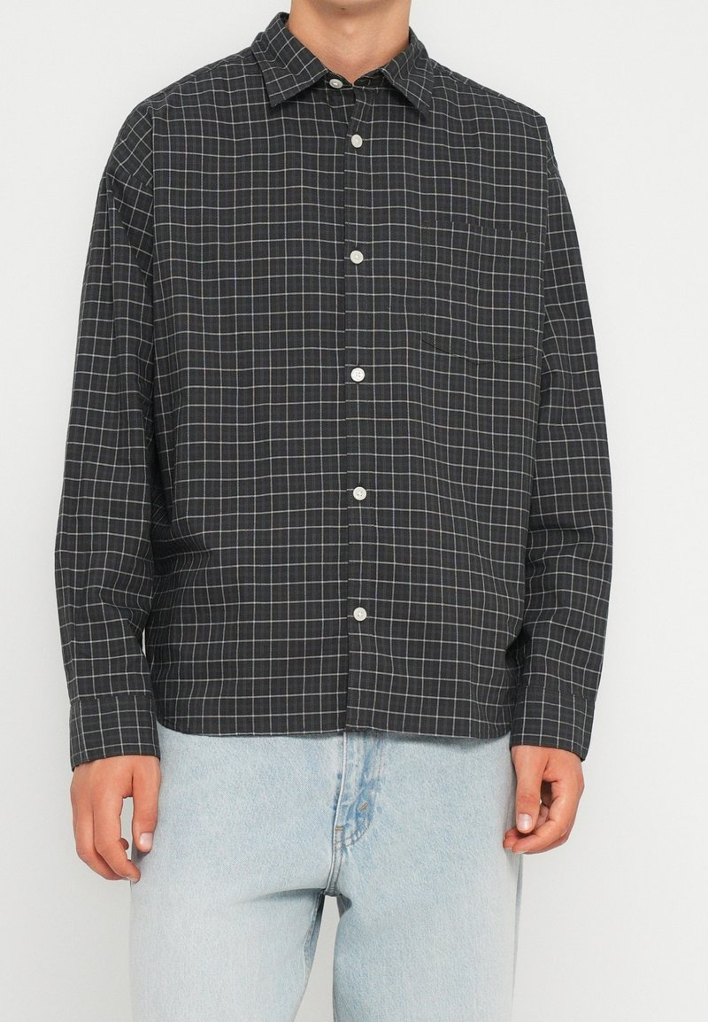 Homme portant une chemise noire à carreaux à manches longues avec des lignes blanches et un jean bleu clair sur fond blanc.