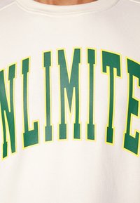 Weißer Sweatshirt mit großem "UNLIMITED"-Text in Grün mit gelber Umrandung. Der Stoff wirkt weich und hat eine strukturierte Oberfläche.