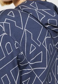 Felpa blu navy con un motivo geometrico bianco, dotata di cappuccio con cordini e tessuto testurizzato per calore e comfort.
