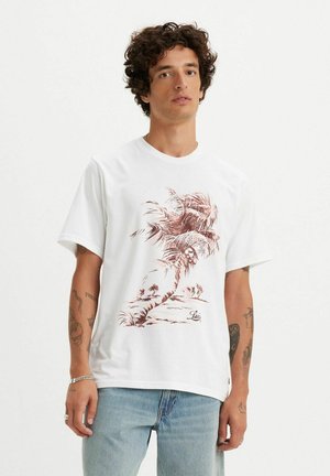 Hvid bomulds t-shirt med et brunt palmetræ grafik og logo. Korte ærmer, rund hals og afslappet pasform, parret med lyseblå jeans.