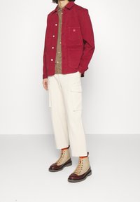 Veste en coton rouge avec des boutons, chemise marron en dessous, pantalon cargo crème et bottes à lacets beiges avec des accents brun foncé.
