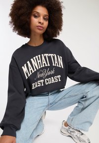 Felpa grigia scura con la scritta "MANHATTAN NEW YORK EAST COAST" in crema; vestibilità oversize, polsini a costine, abbinata a jeans chiari strappati e sneakers argentate.