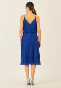 Blaues, gefälteltes Kleid mit V-Ausschnitt, verstellbaren Spaghettiträgern und einem schwingenden Rock. Getragen mit silbernen High-Heels.