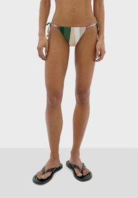 Bikini-Höschen mit grünen, weißen und rostfarbenen Streifen. Bindebänder an den Seiten. Kombiniert mit schwarzen Flip-Flops. Glatte Stofftextur, minimaler Schnitt.