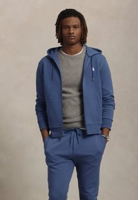 Blå huvtröja med framfickor, i kombination med grå rundhalsad tröja och matchande blå joggers. Avslappnad passform, bomullsmaterial, avslappnad design.