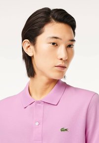 Polotröja i ljusrosa, med klassisk krage och två knappar. Noterbar grön broderad logotyp på vänster bröst. Texturerat tyg.