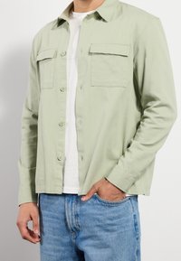 Hombre con una camisa verde claro de botones sobre una camiseta blanca, con jeans azules, mano en el bolsillo, contra un fondo liso.
