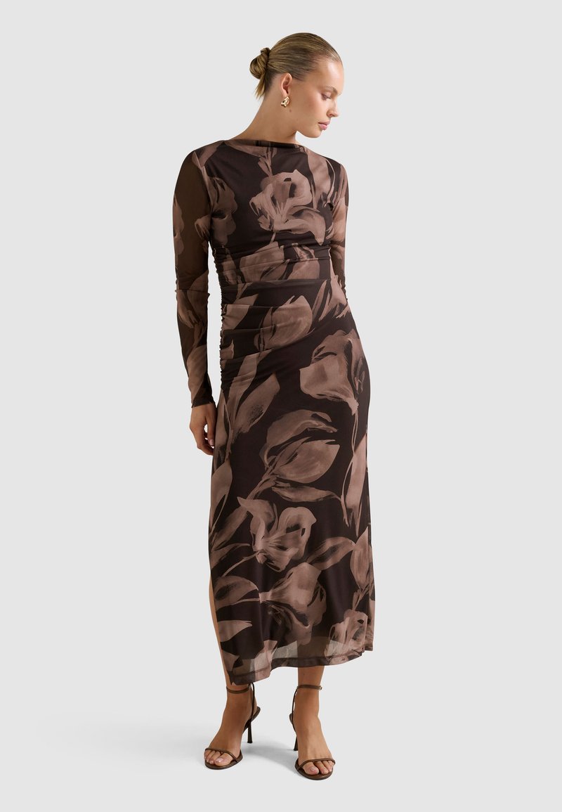 Donna in un lungo vestito aderente a maniche trasparenti di colore marrone con grandi motivi floreali, indossa orecchini d'oro e tacchi con cinturini marroni, in posa di profilo.