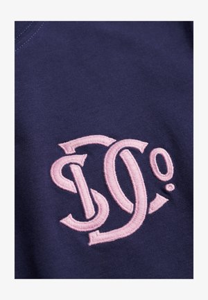 Marineblaue Baumwollstoff mit einem pinkfarbenen, bestickten Logo aus geschwungenen Buchstaben "D" und "O" mit Akzenten und einem Punkt.